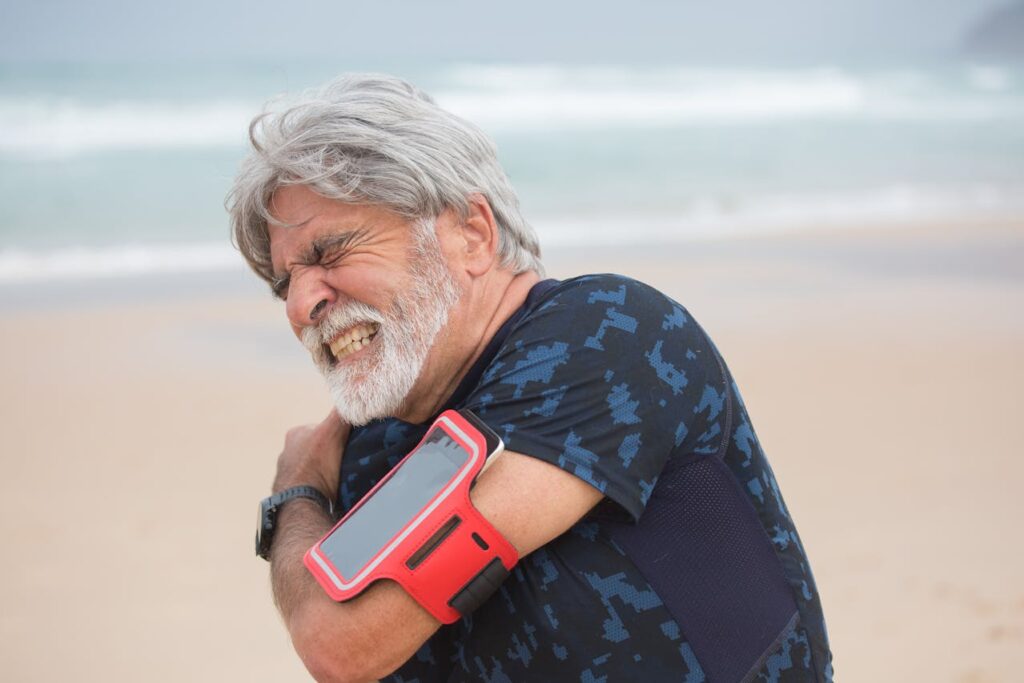 Homme âgé souffrant de douleur à l'épaule avec prothèse, tenant son bras sur une plage.